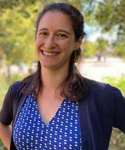 Dr. Greenberg smiles warmly in a blue and white blouse with a navy cardigan in front of the trees lining the boulevard behind the Department of Psychology.