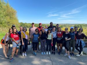 A group of 19 graduate students gather together for a photo under a clear blue sky at a local conservation park.