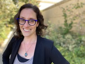 Wearing bright blue glasses, a black blazer, and an aqua pendant, curly-haired Karen Schloss smiles in front of a background of greenery