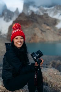 A woman with a red winter hat, black jacket, and black pants holds a Canon camera while stiting on a rock in front of a blurry background of mountains and a lake. 