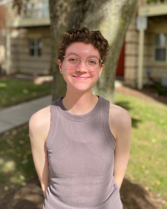 Mia has short brown curly hair, and wears round wire-rimmed glasses. SHe is wearing a light brown tank top and stands smiling in front of a tree. 