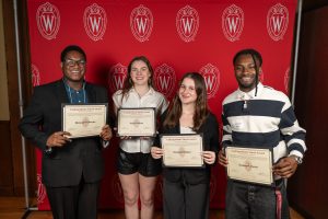 In front of a red backdrop, four students pose with their certificates. 