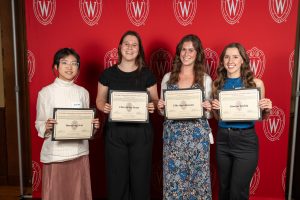 Four recipients pose with their certificates in front of a red backdrop.