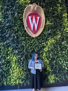 Molly Moen holds her framed award in front of a green wall with a UW crest centered above her.